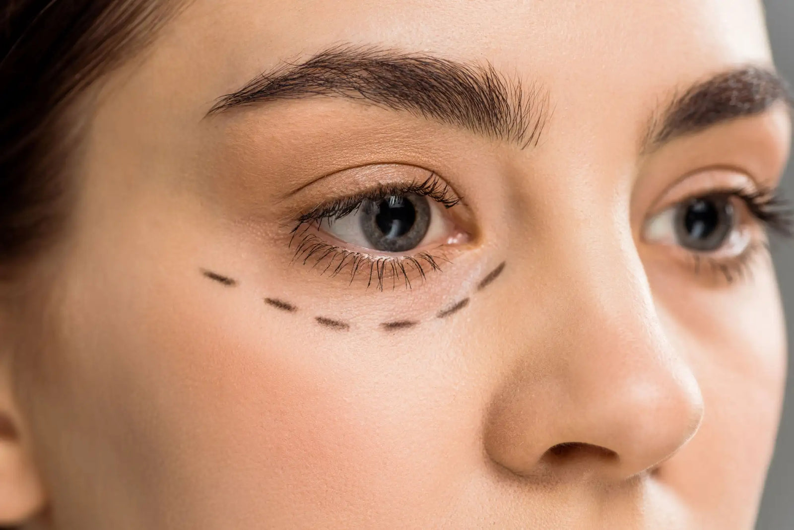 A close-up of a woman’s face with dark brown eyebrows and gray eyes, featuring dashed black lines drawn under her right eye.